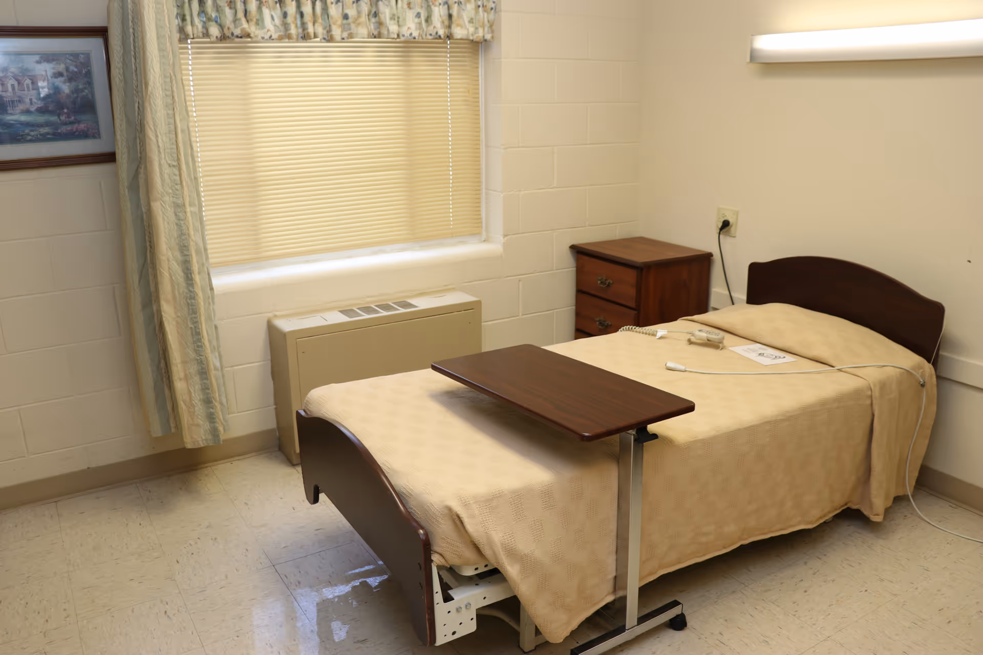 A simple healthcare facility bedroom with a single bed covered in a beige blanket, a wooden overbed table, a small wooden nightstand with drawers, a window with closed blinds and floral curtains, and a framed picture on the wall.