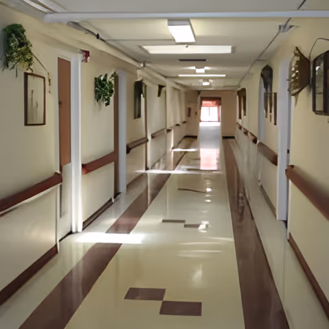 A long, clean hallway inside a senior living facility with beige walls, brown handrails on both sides, several closed doors, framed mirrors, and small green plants mounted on the walls. The floor has a polished pattern with light and dark tiles, and fluorescent ceiling lights illuminate the corridor.