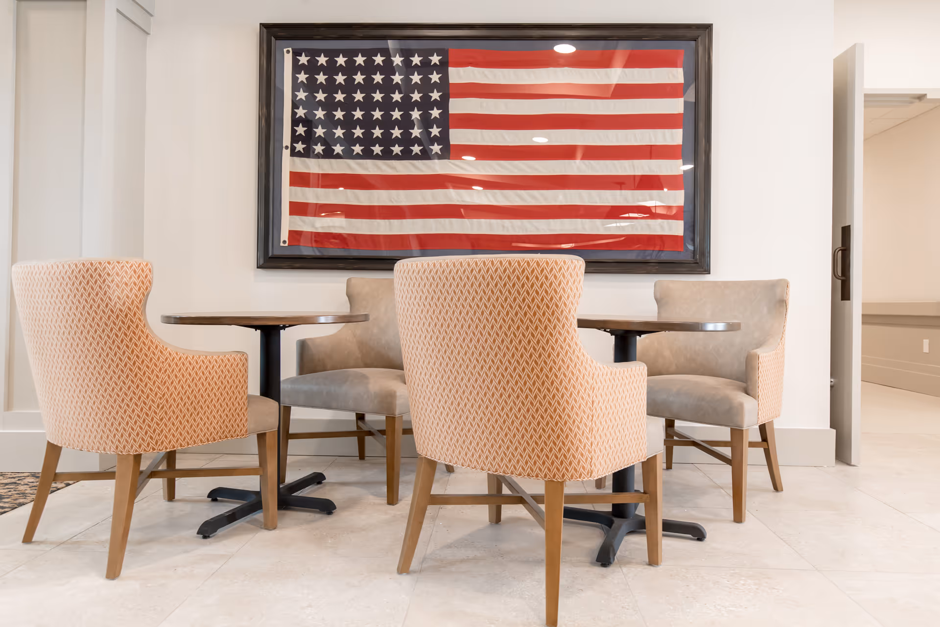 A seating area with four upholstered chairs around two small round tables, positioned in front of a large framed American flag mounted on a white wall. The floor is tiled, and there is an open doorway to the right.