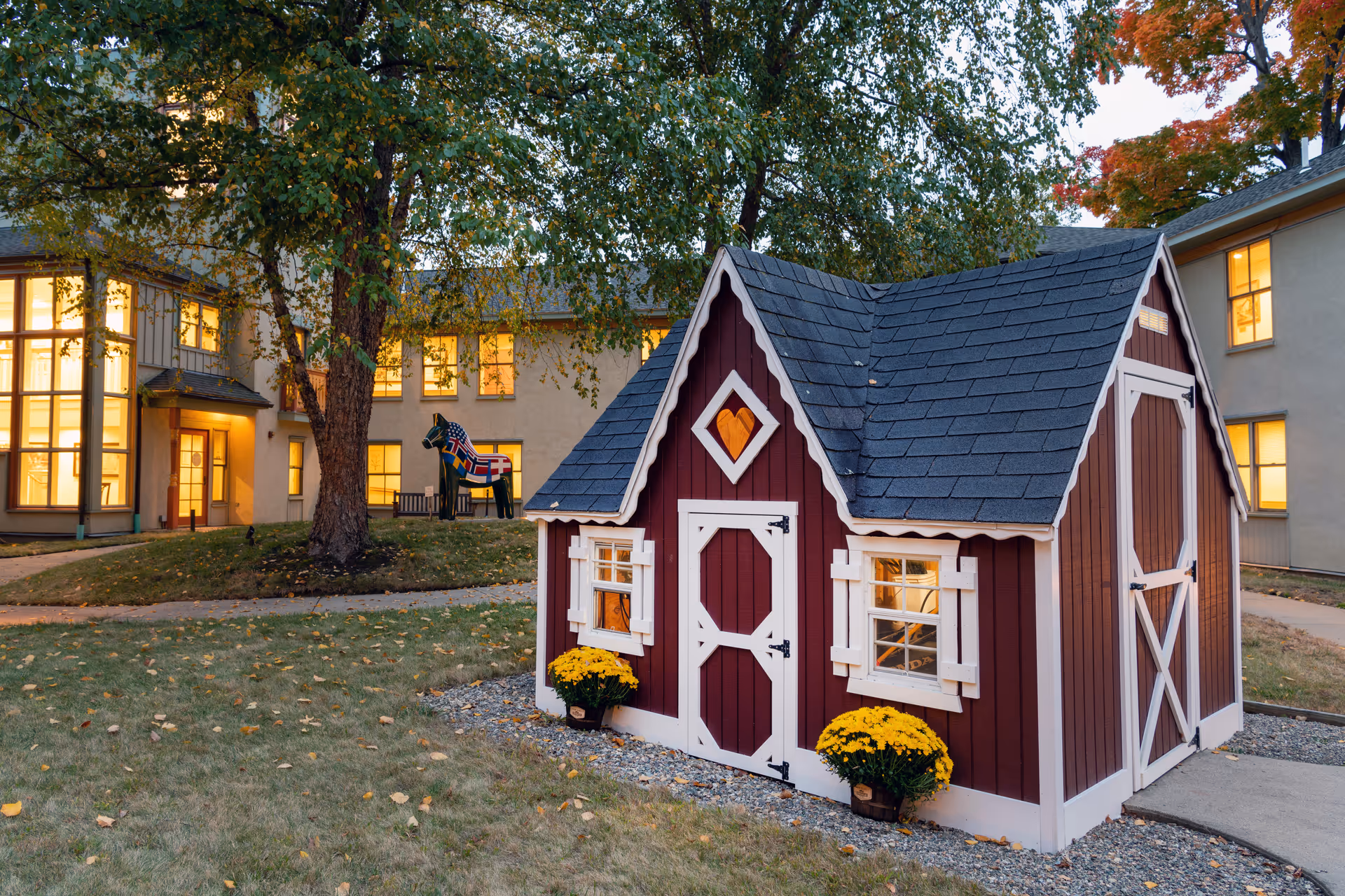 A small decorative red and white playhouse with a heart-shaped cutout above the door, surrounded by yellow flowers and situated on a grassy area with a large tree nearby. In the background, there is a building with many lit windows and a wooden horse painted with Scandinavian flags.