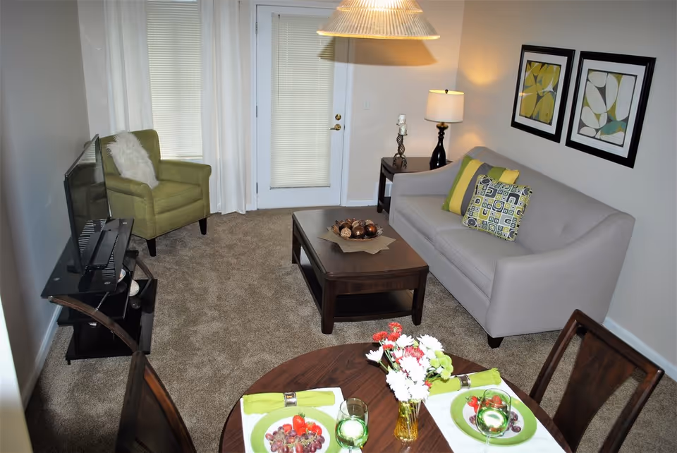 Cozy furnished living room with a gray sofa, green armchair, coffee table, TV on a stand, and a small dining table set with plates and flowers.