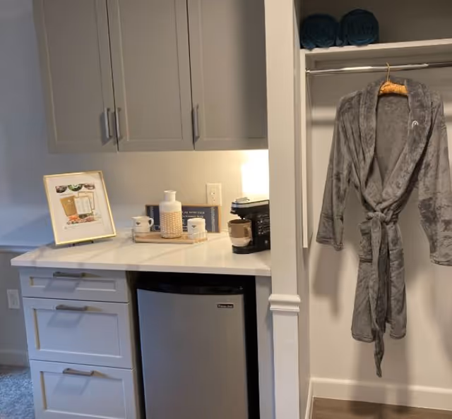 Small kitchenette with upper cabinets, a countertop, mini fridge and coffee items next to an open closet with a hanging gray robe.