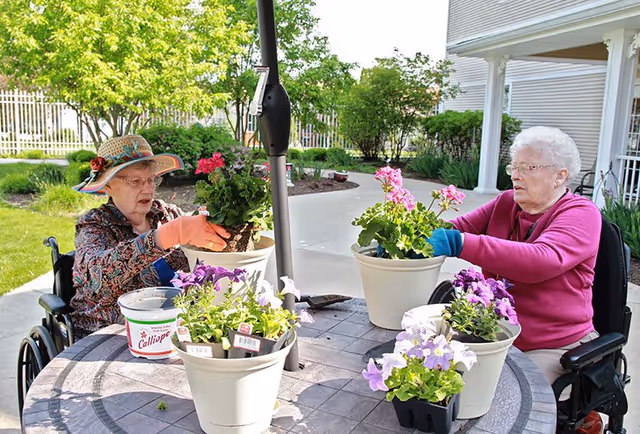 Two elderly women in wheelchairs are gardening outdoors at a round table with several flower pots containing colorful flowers. One woman is wearing a floral patterned jacket and a hat, while the other is wearing a pink sweater and glasses. They are both wearing gardening gloves and appear to be enjoying the activity in a sunny garden area with green grass and bushes in the background.