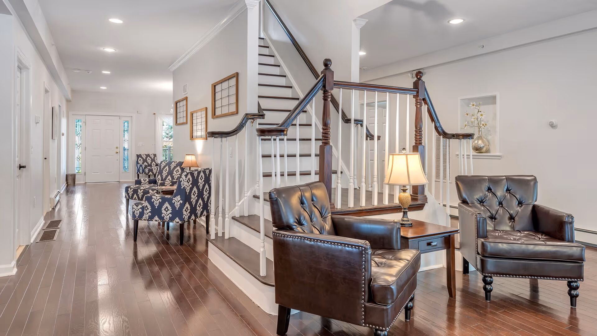 A bright and spacious interior area of Clover Hill Senior Living featuring a wooden staircase with white railings and dark wood banisters. There are two dark leather armchairs with a wooden side table and lamp between them in the foreground. Further back, two patterned armchairs with a small table and lamp are placed along the hallway leading to a white front door with glass panels on either side. The floor is polished dark wood, and the walls are painted light gray with some framed artwork and decorative items.
