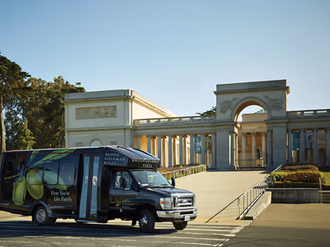 A black shuttle van with Rhoda Goldman Plaza branding is parked on a street in front of a large classical-style building with columns and an archway under a clear blue sky.