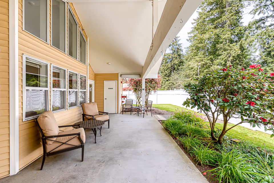Covered outdoor patio area with cushioned chairs and small tables along the side of a yellow building. There are flowering bushes and greenery next to the patio, with a white fence and tall trees in the background.