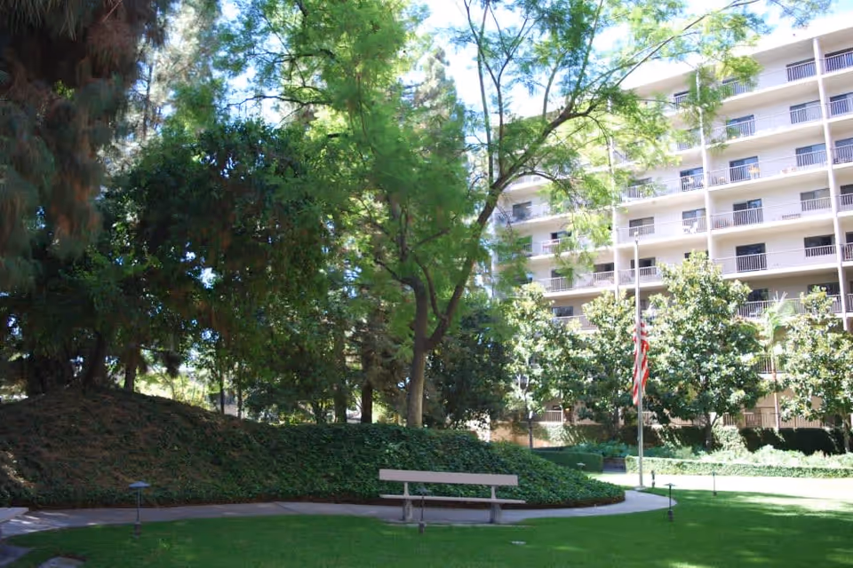 Outdoor garden area with a green lawn, a curved pathway, a bench, several trees, and a multi-story building with balconies in the background. An American flag is also visible near the center of the image.