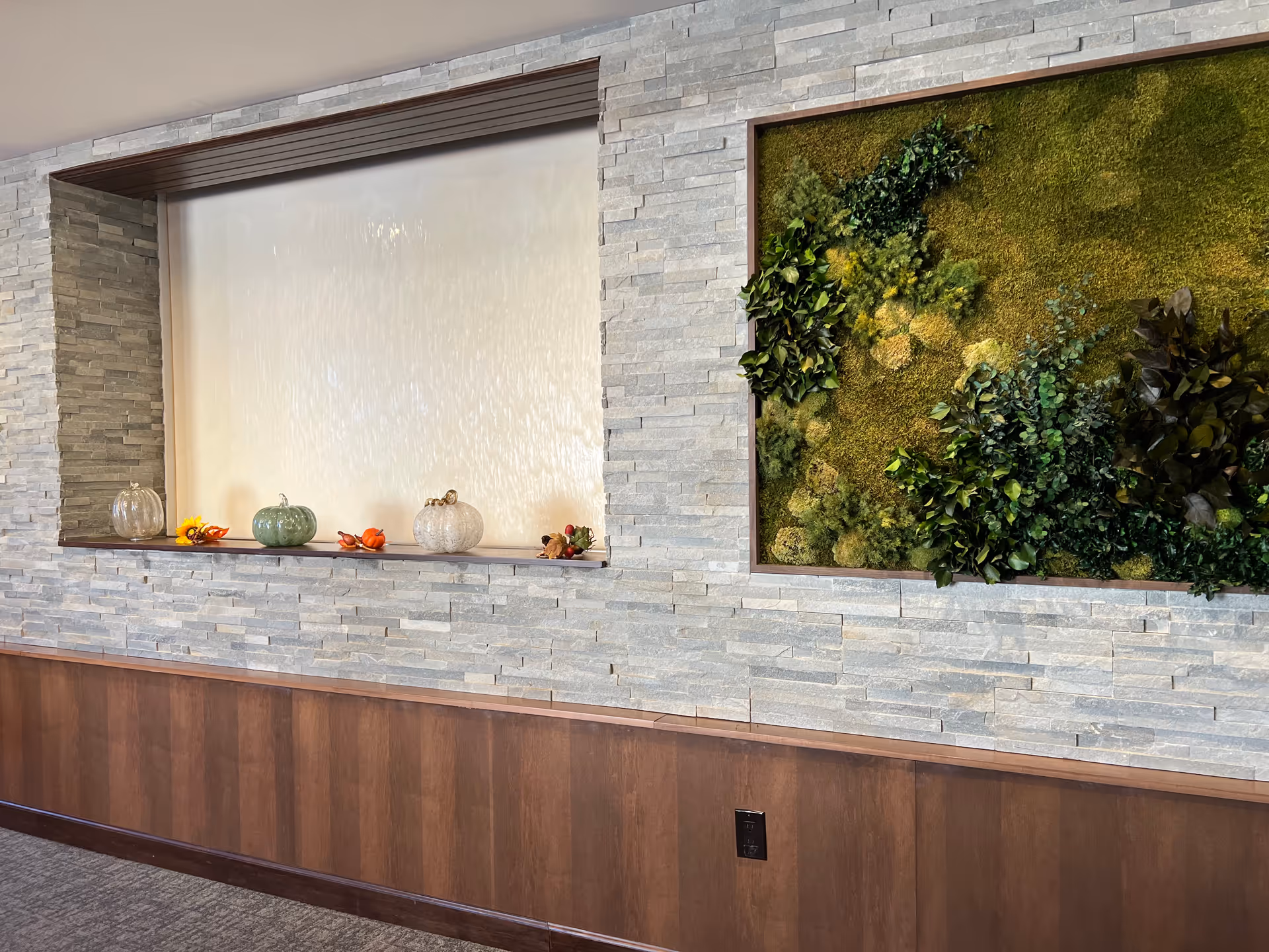 Interior wall with light gray stone tiles and dark wood paneling at the bottom. The wall features a recessed water feature with a smooth white surface and decorative small pumpkins and fall-themed items on the ledge. Next to it is a framed green moss and plant wall art installation.