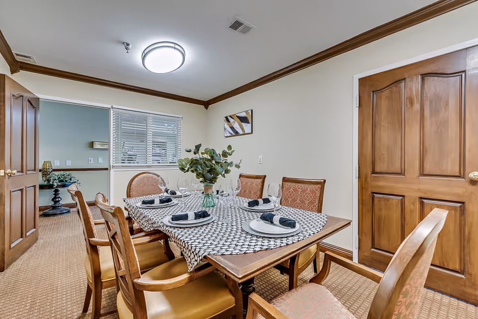A dining room with a rectangular wooden table set for six people. The table has a patterned tablecloth, white plates with black napkins, wine glasses, and a green vase with leafy plants as a centerpiece. The room has beige walls, a wooden door, a window with white blinds, and a ceiling light fixture. There is a small piece of abstract art on the wall and an open doorway leading to another room with a small table and plants.
