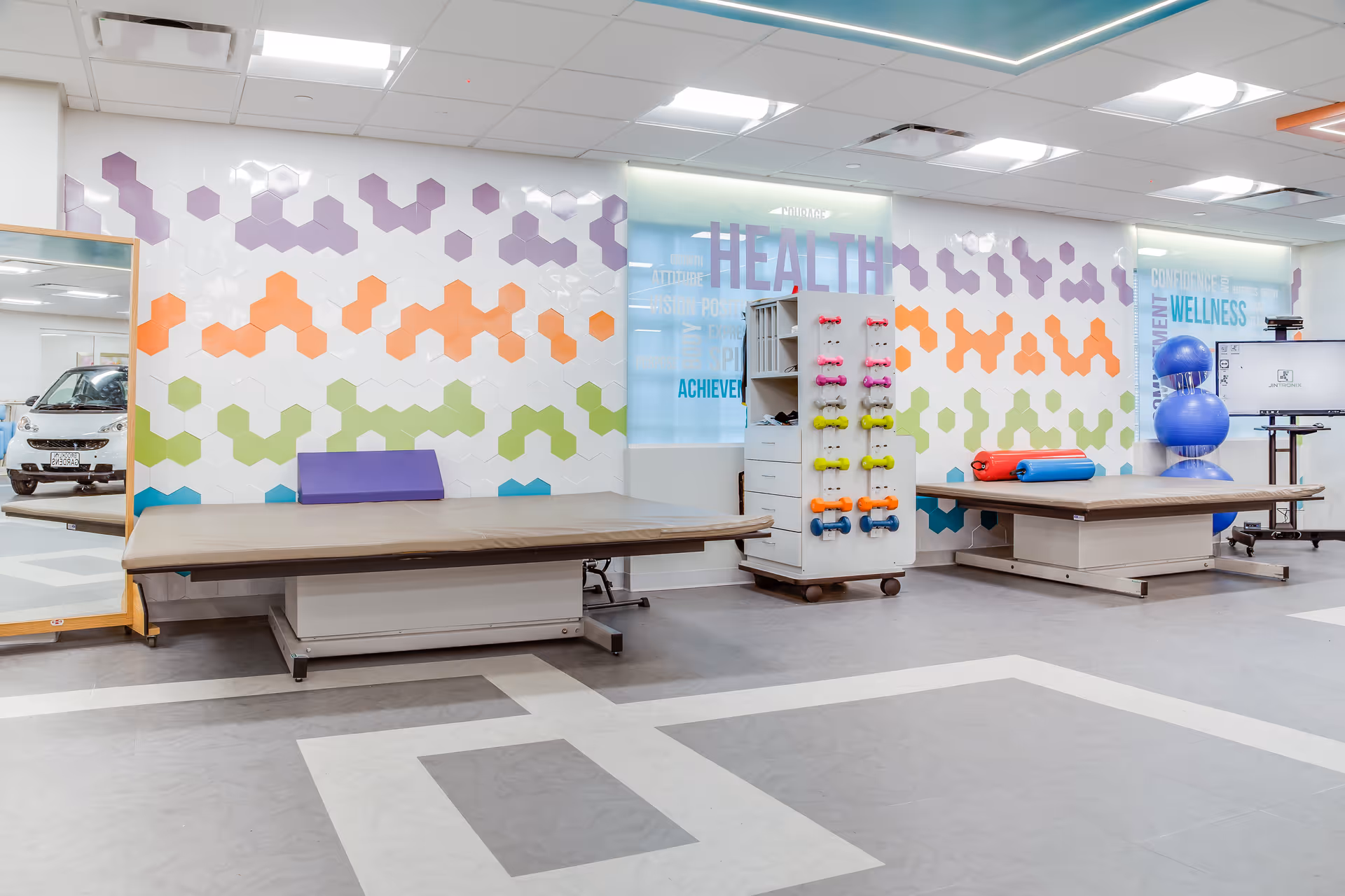 A bright and clean rehabilitation room with two large padded therapy tables, a rack holding colorful small dumbbells, blue exercise balls stacked in the corner, and a large mirror reflecting part of the room. The walls are decorated with colorful hexagonal patterns and motivational words like 'HEALTH' and 'WELLNESS'.