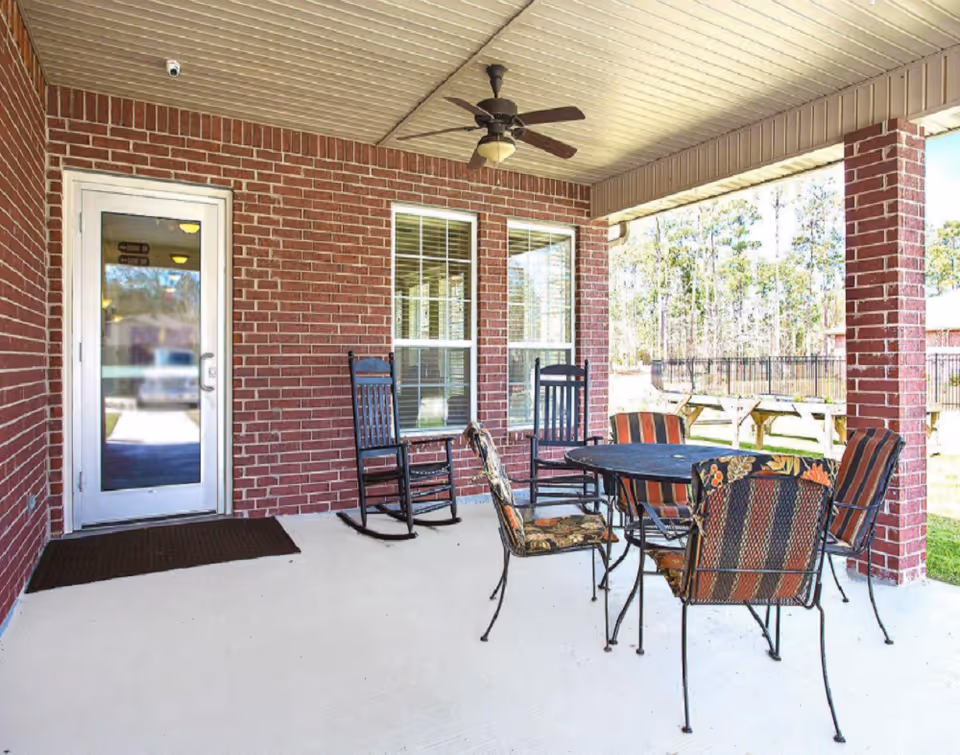 Covered brick patio with a round table, patterned metal chairs, two rocking chairs, and a ceiling fan.