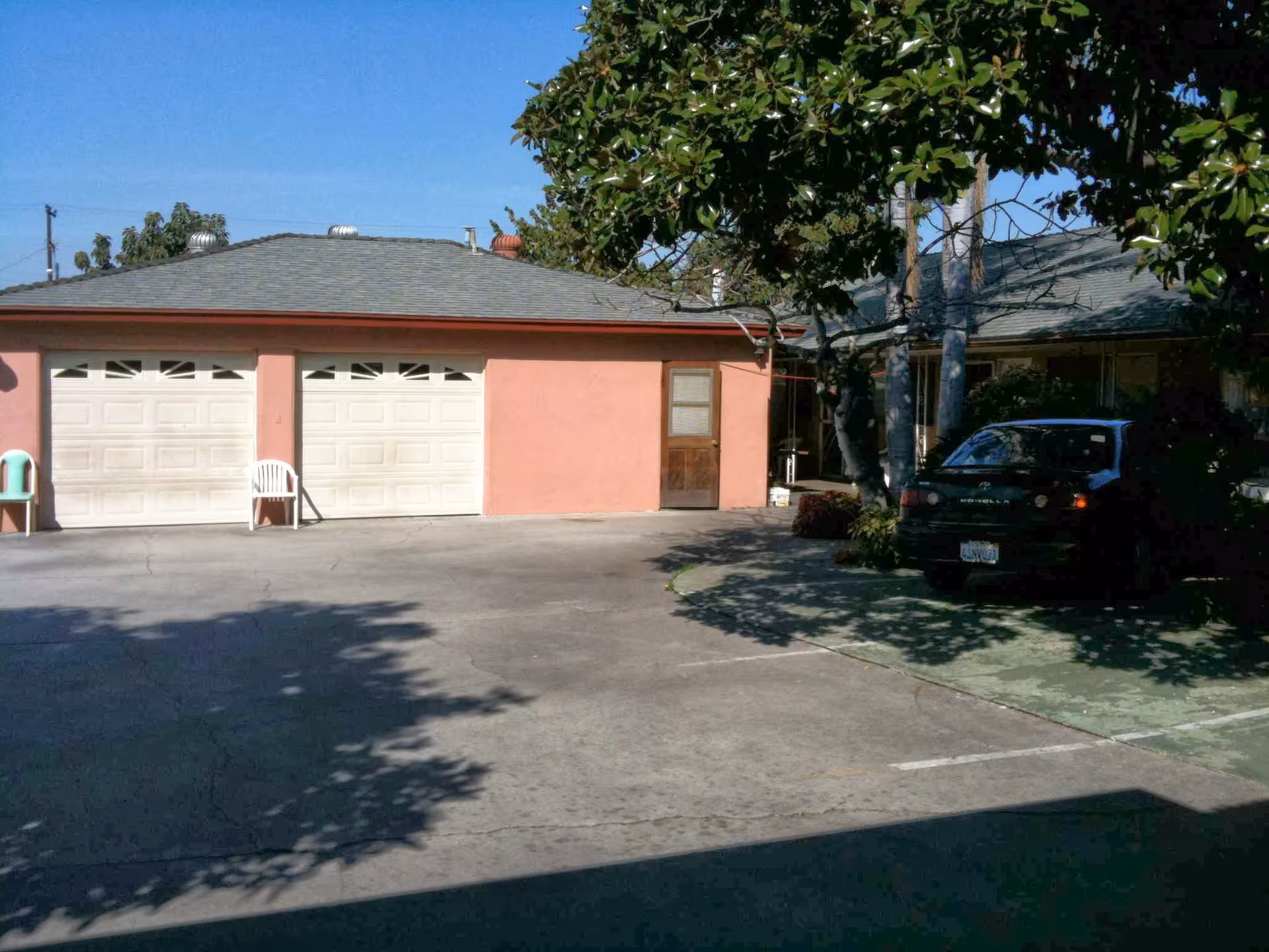 Exterior view of a residential building with a double garage door, a single door, two white plastic chairs, a parked black car, and trees casting shadows on the driveway under a clear blue sky.