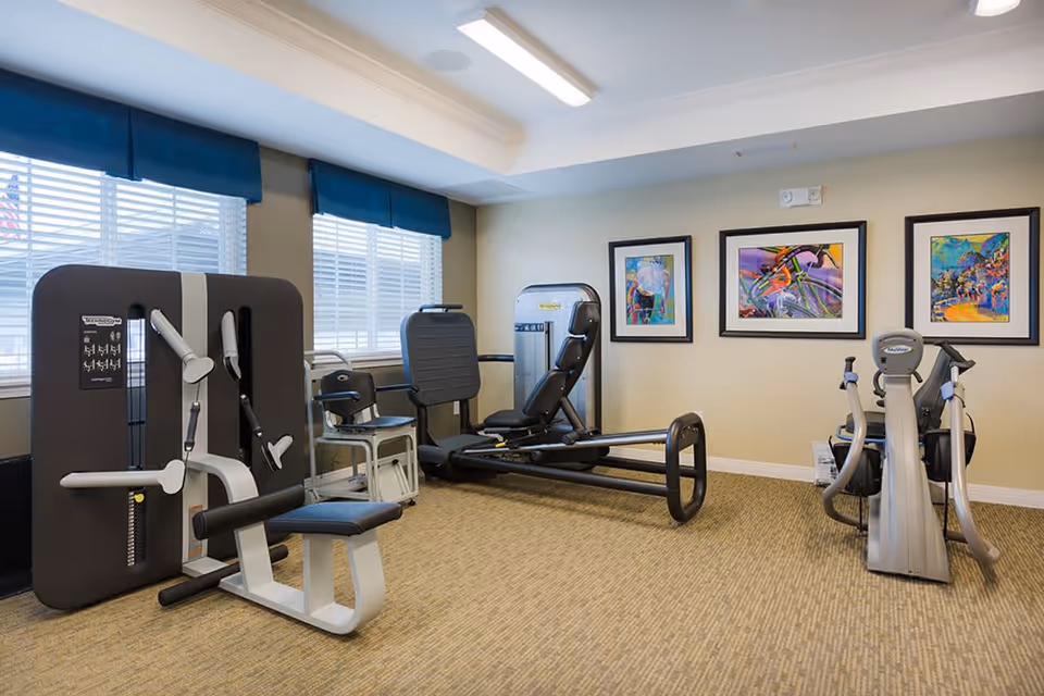 A small fitness room with exercise equipment including a seated leg press machine, a rowing machine, and a stationary bike. The room has beige walls, carpeted floor, three framed colorful artworks on the wall, and two windows with blue valances letting in natural light.
