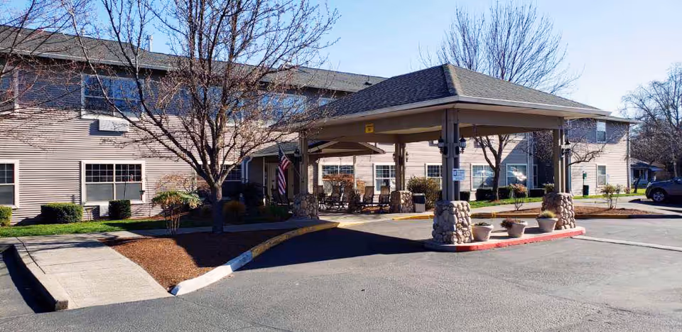 Exterior view of a two-story senior living facility building with a covered entrance supported by stone pillars. There are leafless trees and some landscaping around the building, with a paved driveway and parking area in front.