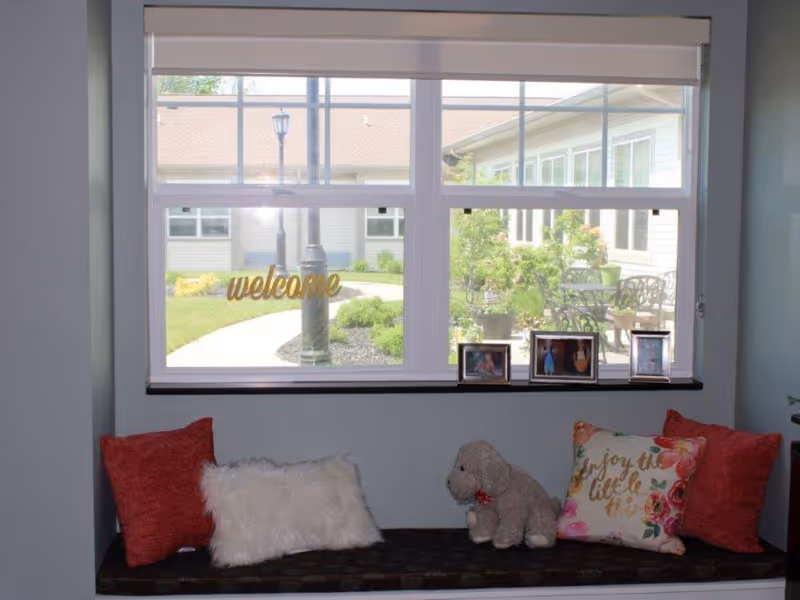 A cozy window seat with red and floral pillows, a white fluffy pillow, and a stuffed dog toy. The window behind shows a view of a courtyard with a lamp post, greenery, and outdoor seating. There are three framed photos on the window sill and a 'welcome' sign on the window glass.
