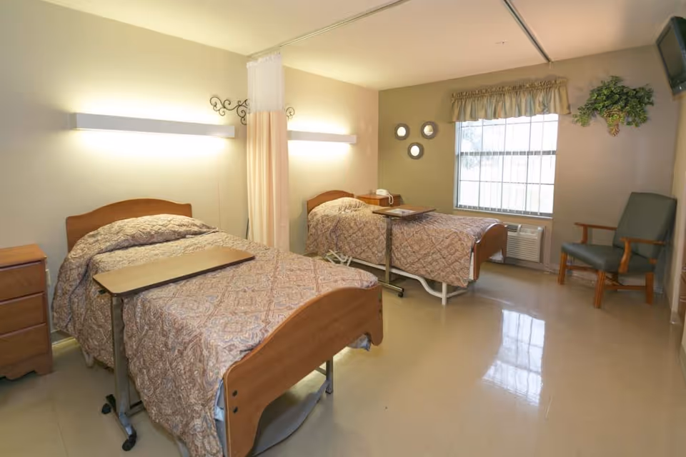 A room with two hospital-style beds, each with a patterned bedspread and an overbed table. The room has a window with a valance, a green armchair, a small wooden nightstand, and wall-mounted lights above each bed. There is a curtain divider between the beds and a wall-mounted TV in the corner.