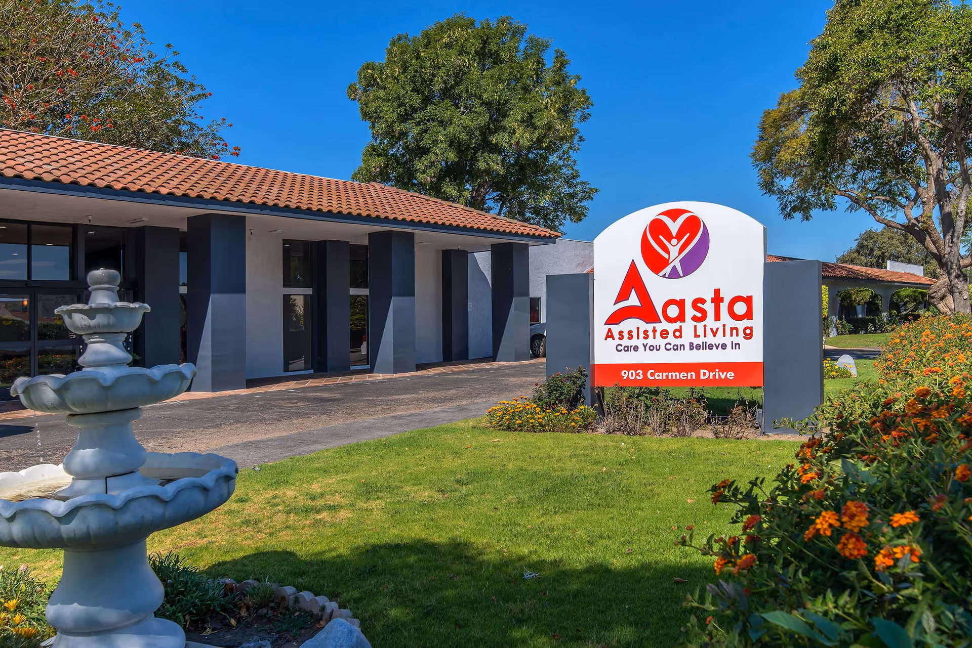 Exterior view of Aasta Assisted Living of Camarillo facility showing a white and red sign with the facility's name and address 903 Carmen Drive, a three-tiered white fountain, green lawn, flowering plants, and trees under a clear blue sky.