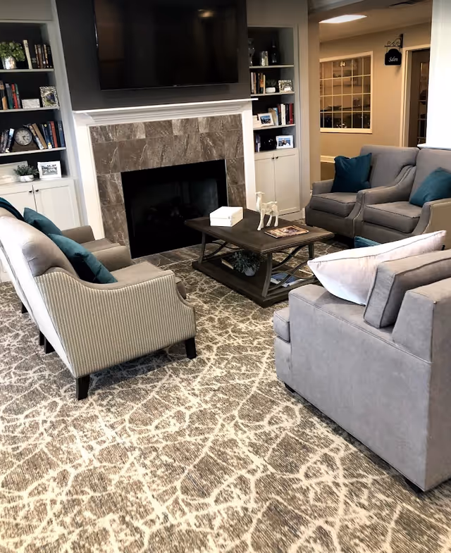 A cozy living room with a patterned carpet, four gray upholstered chairs with teal and white pillows arranged around a dark wooden coffee table. The coffee table has a small decorative white horse and a book on it. Behind the seating area is a fireplace with a tiled surround and a large flat-screen TV mounted above it. Built-in shelves with books and decorative items flank the fireplace. The room has soft lighting and a neutral color palette.