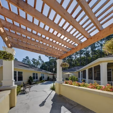 Outdoor patio area at The Addison of Bluffton featuring a wooden pergola overhead, benches along the walkway, hanging flower baskets, and surrounding buildings with a garden bed filled with flowers. Trees are visible in the background under a clear blue sky.