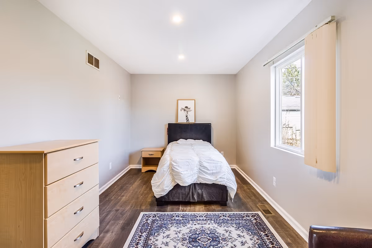A small minimalist bedroom with a single bed centered against the far wall, a wooden dresser and nightstand, a window on the right, and a patterned rug on dark wood flooring.