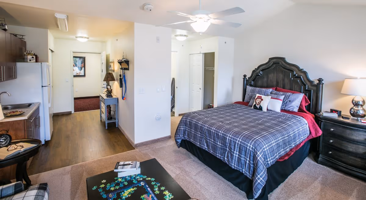 A cozy studio apartment featuring a bed with a dark wooden headboard and plaid bedding, a nightstand with a lamp and books, a small kitchenette with a sink, refrigerator, and cabinets, and a coffee table with a partially completed puzzle. The room has a ceiling fan with a light, carpeted and wooden flooring, and a hallway leading to other areas.