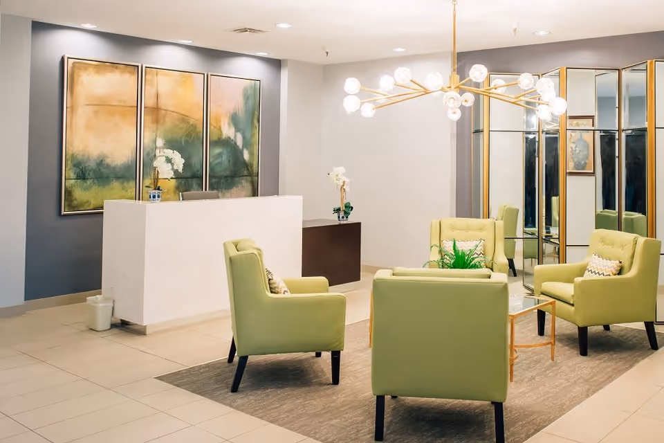 A bright reception lounge with green armchairs around a glass coffee table, a white reception desk, abstract wall art and a modern chandelier.