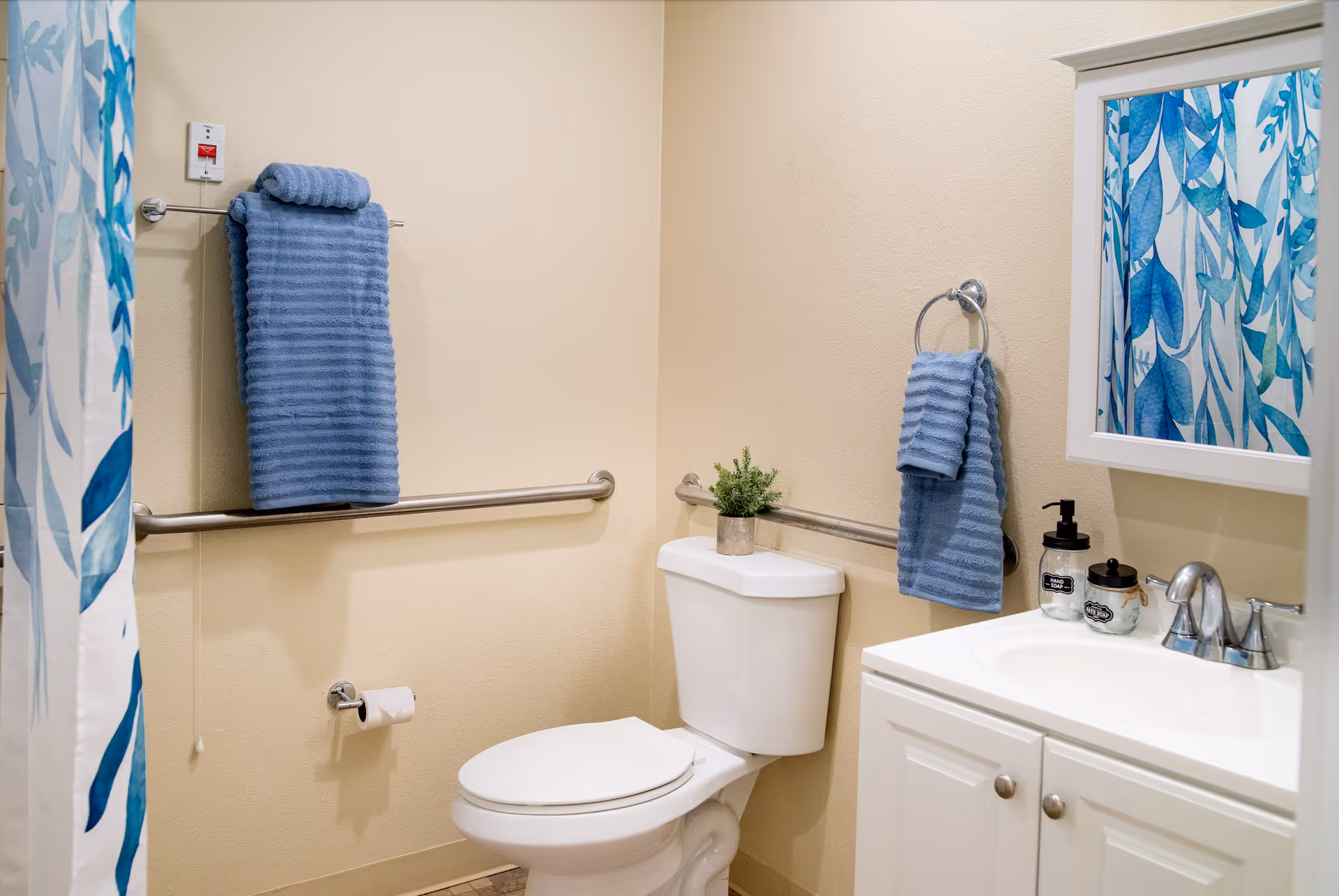 A clean bathroom with a white toilet and a white vanity with a sink. Blue towels hang on a towel bar and a ring, and a small green plant sits on the toilet tank. A shower curtain with blue and green leaf patterns is partially visible. The walls are beige, and there are grab bars installed near the toilet and towel bar.