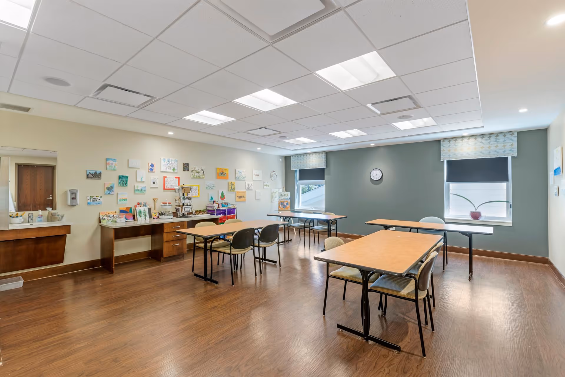 A well-lit activity room with wooden floors and several tables and chairs arranged for group activities. The walls are decorated with various colorful artworks and crafts. There are two windows with patterned valances and black roller shades, a clock on the wall, and a counter area with supplies and a sink.