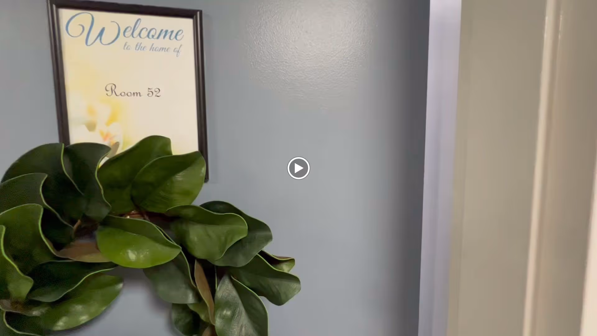 Framed 'Welcome' sign for Room 52 on a light blue interior wall with a large green leafy wreath beneath it.