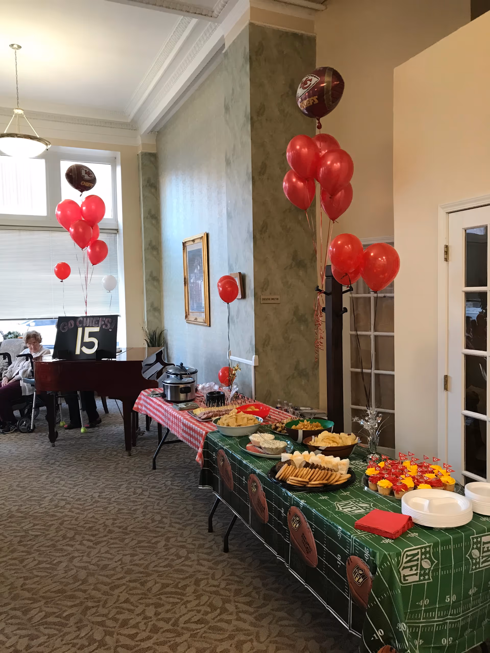 A decorated indoor space with tables covered in red and green tablecloths featuring football-themed designs. The tables hold various snacks and cupcakes with small flags. Red and white balloons, including some football-shaped ones, are tied to a coat rack and other spots. In the background, a piano with a sign reading 'GO CHIEFS 15' is visible, along with two seated elderly women near a large window.