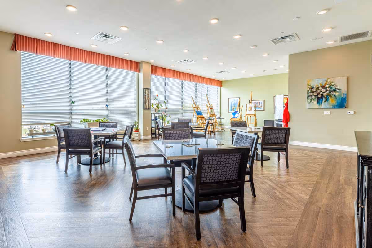 A bright and spacious room with large windows covered by blinds and red valances. The room contains several square tables surrounded by chairs with patterned backs and cushioned seats. In the background, there are easels with artwork displayed, potted plants near the windows, and framed pictures on the walls. The floor is wood-patterned, and the ceiling has multiple recessed lights.