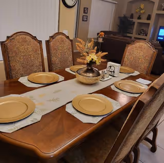 A wooden dining table set with six ornate chairs, each with a patterned fabric backrest. The table has a beige table runner with a floral design, six gold-colored plates on placemats, and a decorative vase with dried flowers in the center. In the background, there is a living room area with a couch, shelves with decorations, and a television.