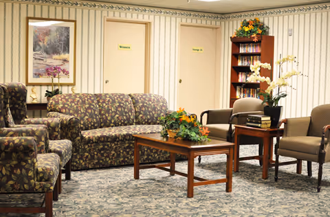 A cozy lounge with patterned sofas and armchairs arranged around a wooden coffee table and side tables in a senior living facility.
