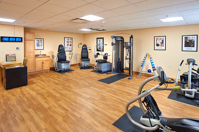 A fitness room with wooden flooring and beige walls featuring various exercise equipment including stationary bikes, weight machines, and a rack of colorful dumbbells. The room has a digital clock on the wall, framed motivational posters, and a small sink and cabinet area in the corner.