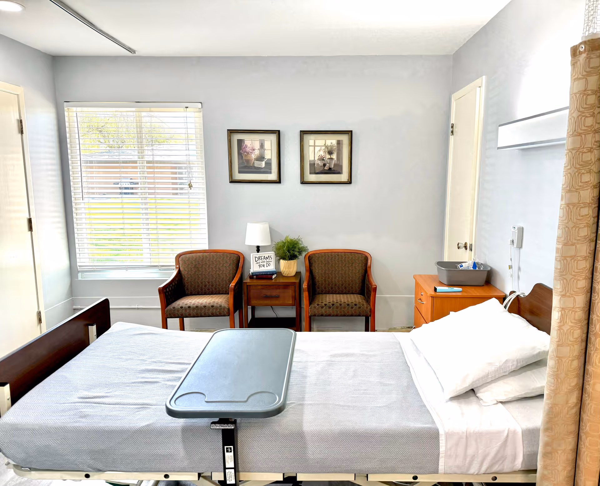 A bright and clean patient room in a senior living facility with a hospital bed covered in white linens and a gray overbed table. Two patterned armchairs flank a wooden side table with a lamp, a small plant, and a sign that reads 'DREAMS don't work unless you DO'. Two framed floral pictures hang on the light gray wall above the chairs. A window with white blinds lets in natural light, and a wooden cabinet with a gray tray is next to the bed. A beige privacy curtain is partially visible on the right side.
