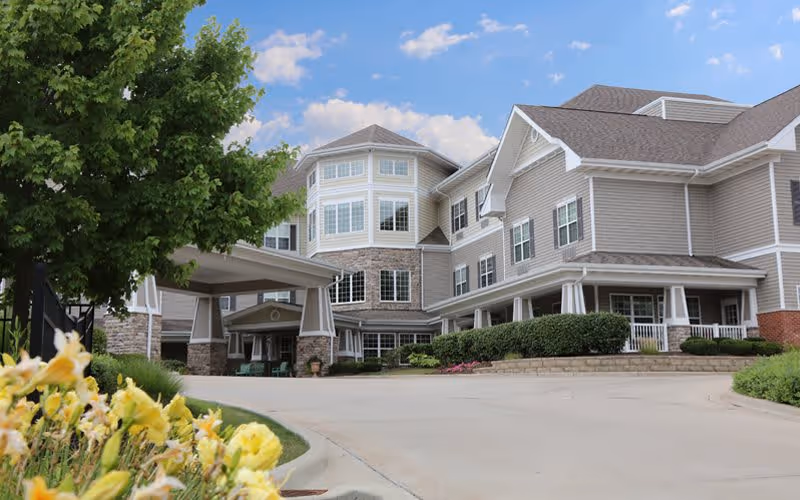 Exterior view of a multi-story senior living facility building with beige siding, white trim, and stone accents. The entrance features a covered drop-off area. There are green bushes and yellow flowers in the foreground, with a clear blue sky and some clouds above.