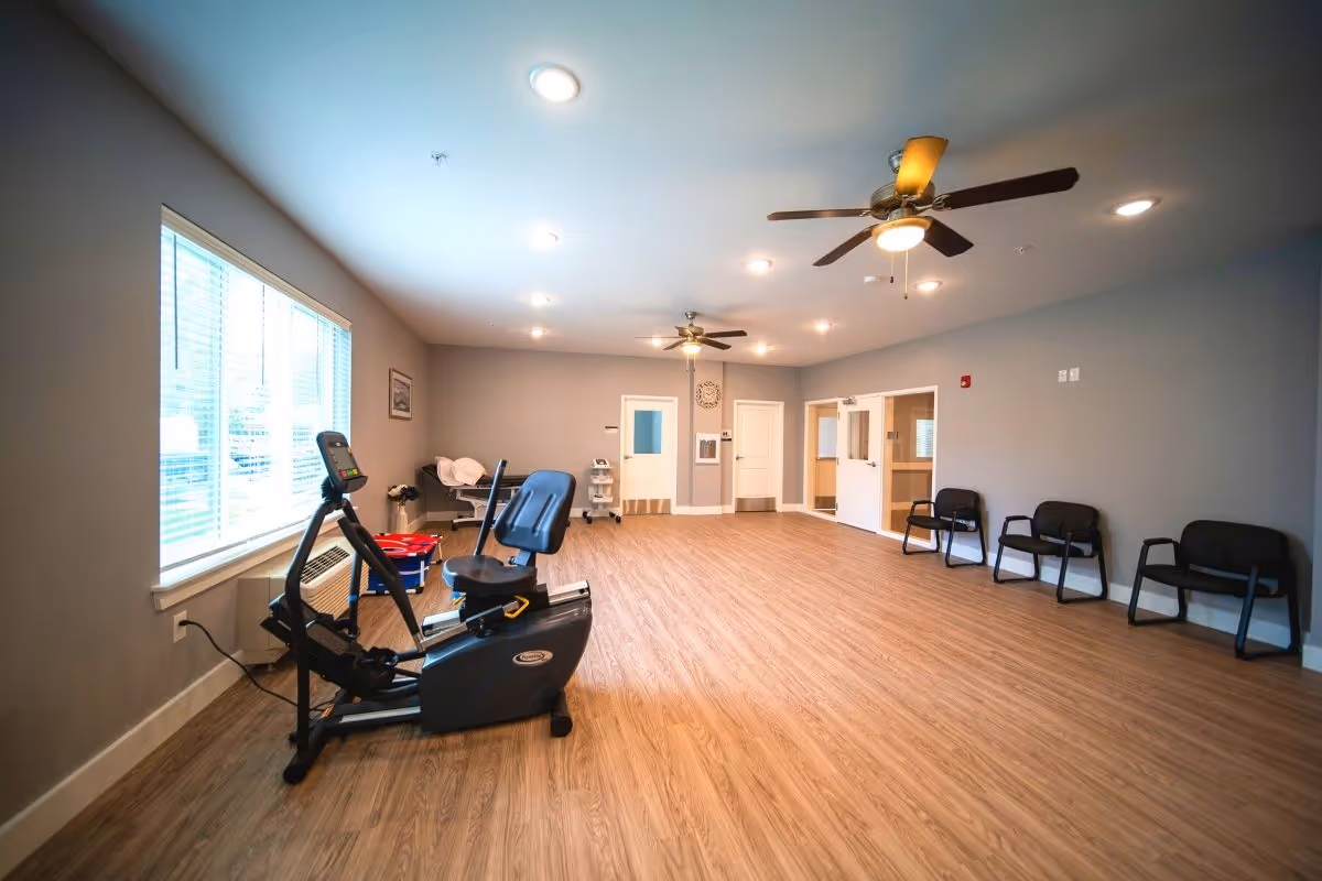 A spacious room with wooden flooring and gray walls, featuring a recumbent exercise bike near a large window with blinds. The room has ceiling fans with lights, several black chairs lined up against the wall, and medical equipment in the corner. The room is well-lit with recessed ceiling lights.