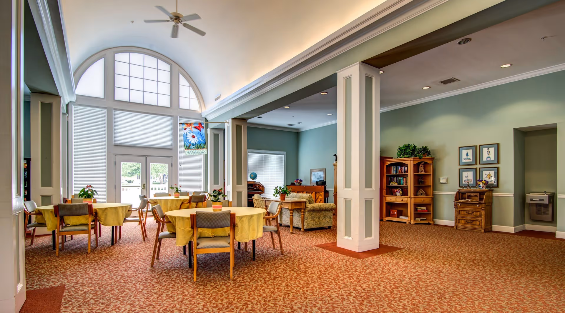Spacious sunny common room with round dining tables and chairs, lounge seating, tall arched windows and decorative columns.