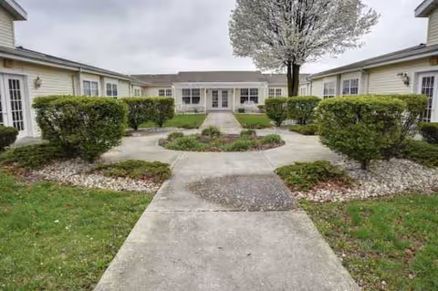 A landscaped courtyard with trimmed hedges, a flowering tree, and a walkway to a single-story building.