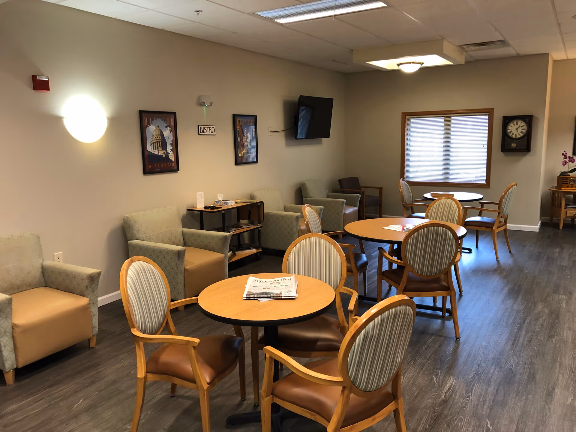 A cozy common area with round wooden tables and cushioned chairs arranged for seating. There are armchairs along the wall, a wall-mounted TV, framed pictures, a clock, and a window with blinds. The floor is wood-style, and the lighting is warm and inviting.