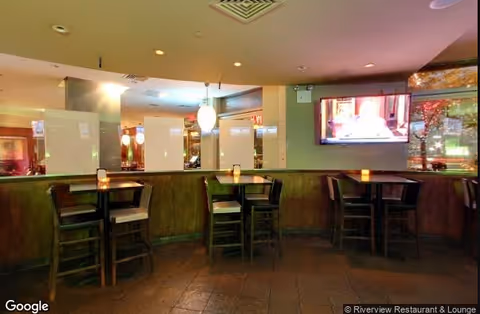 Interior view of a dining area with several small tables and chairs arranged along a curved wooden partition. There are hanging pendant lights above the tables and a flat-screen TV mounted on the wall displaying a program. The floor is tiled, and the atmosphere is softly lit.