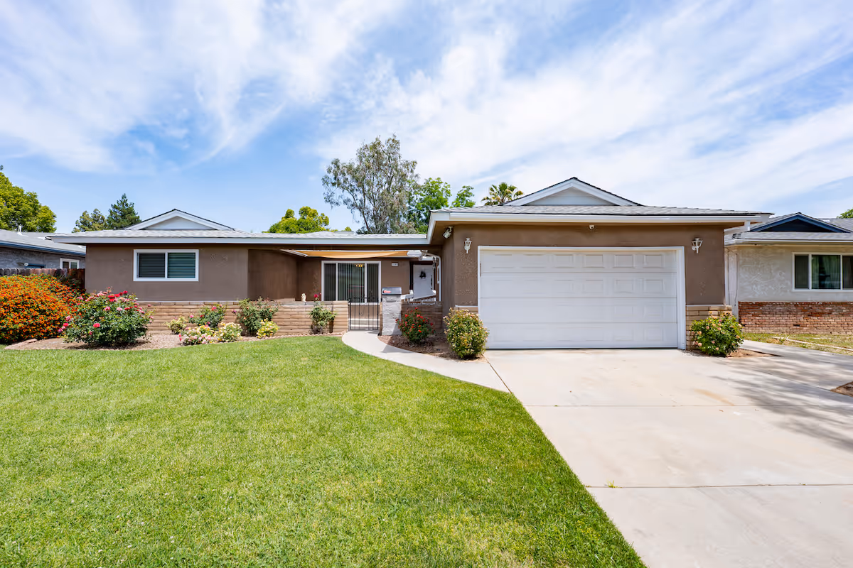 Single-story residential building with a two-car garage, brown exterior walls, a well-maintained green lawn, and flower bushes in front under a partly cloudy sky.