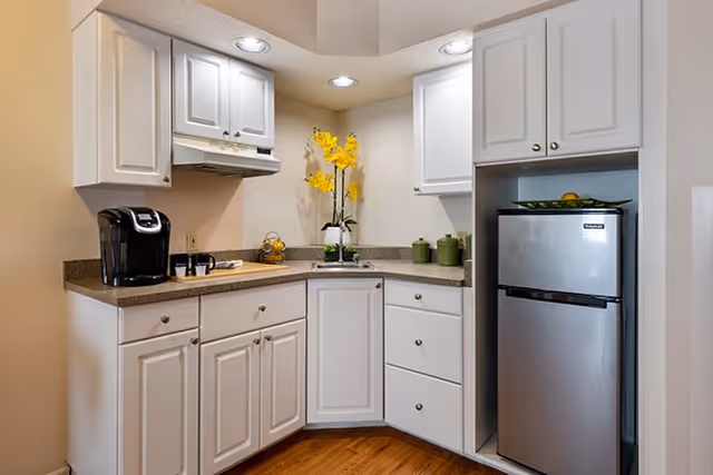 A small kitchen area with white cabinets, a countertop with a coffee maker, cups, a bowl of lemons, and a vase with yellow flowers. There is a compact stainless steel refrigerator and recessed ceiling lights illuminating the space.