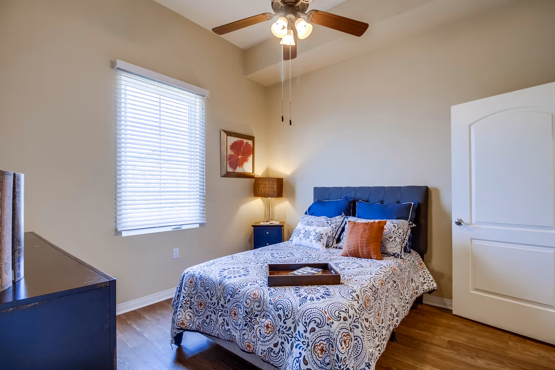 A bedroom with a bed covered in a patterned bedspread and multiple pillows, a wooden tray on the bed, a nightstand with a lamp and a framed flower picture on the wall, a window with blinds, a ceiling fan with lights, and a closed white door.