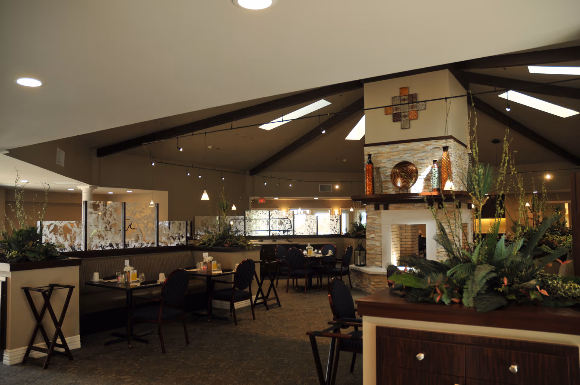 Interior view of a dining area in a senior living facility with tables and chairs arranged around a central fireplace. The space features a high ceiling with exposed beams, pendant lighting, decorative plants, and frosted glass partitions.