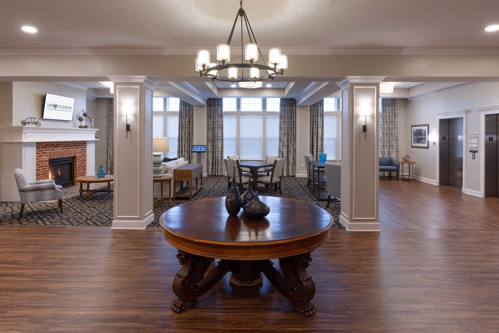Spacious and well-lit common area in The Culpeper facility featuring a round wooden table with decorative vases in the foreground, a chandelier overhead, a seating area with chairs and a fireplace on the left, large windows with curtains in the background, and two elevators on the right side.
