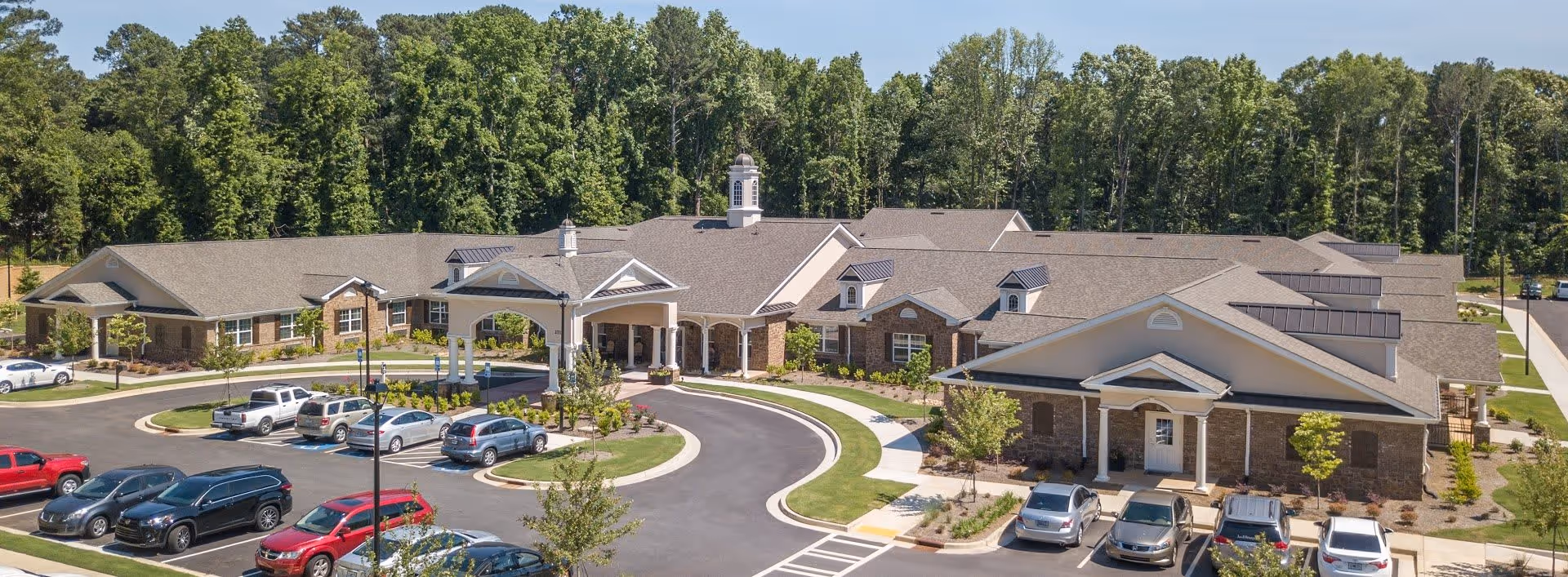 Aerial view of Benton House of Decatur senior living facility showing a large single-story building with a covered entrance, surrounded by a parking lot with several cars and landscaped greenery, with a backdrop of tall trees under a clear blue sky.
