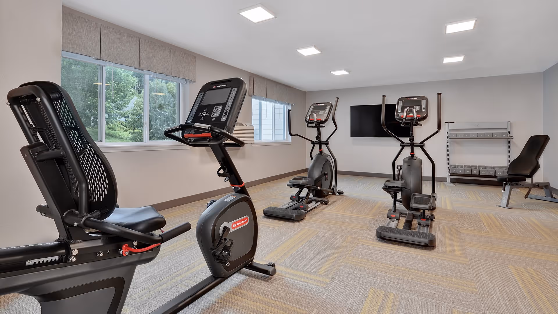 A fitness room with exercise equipment including a recumbent bike, two elliptical machines, a weight bench, and a rack of dumbbells. The room has large windows with beige valances, light-colored walls, and a wall-mounted flat screen TV.