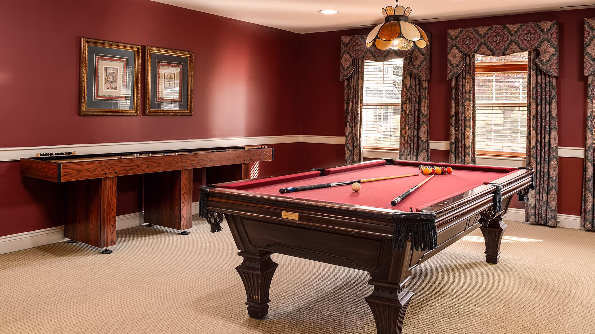 A game room with a red pool table set up for a game, including pool cues and balls. The room has deep red walls, beige carpet, three windows with floral curtains, and a wooden shuffleboard table against the wall. Two framed pictures hang above the shuffleboard table.