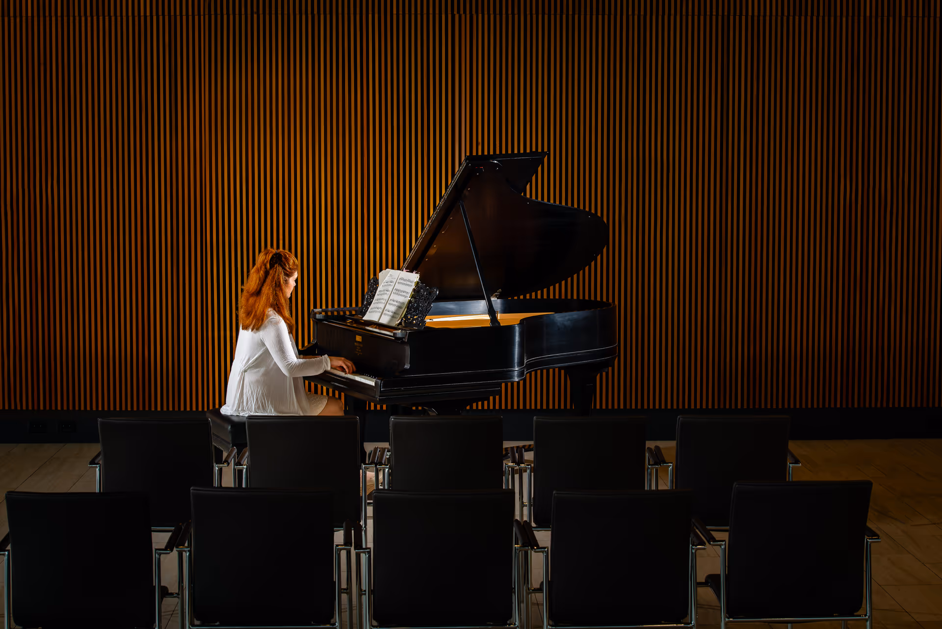 A woman in a white dress plays a grand piano on a small stage with rows of chairs and a wooden slat wall behind her.